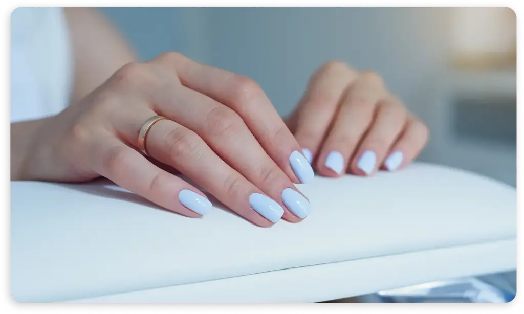Minimalist güzellik ortamında temiz beyaz yüzeyde nazikçe dinlenen yumuşak pastel mavi manikürlü kadın ellerinin yakın çekimi.